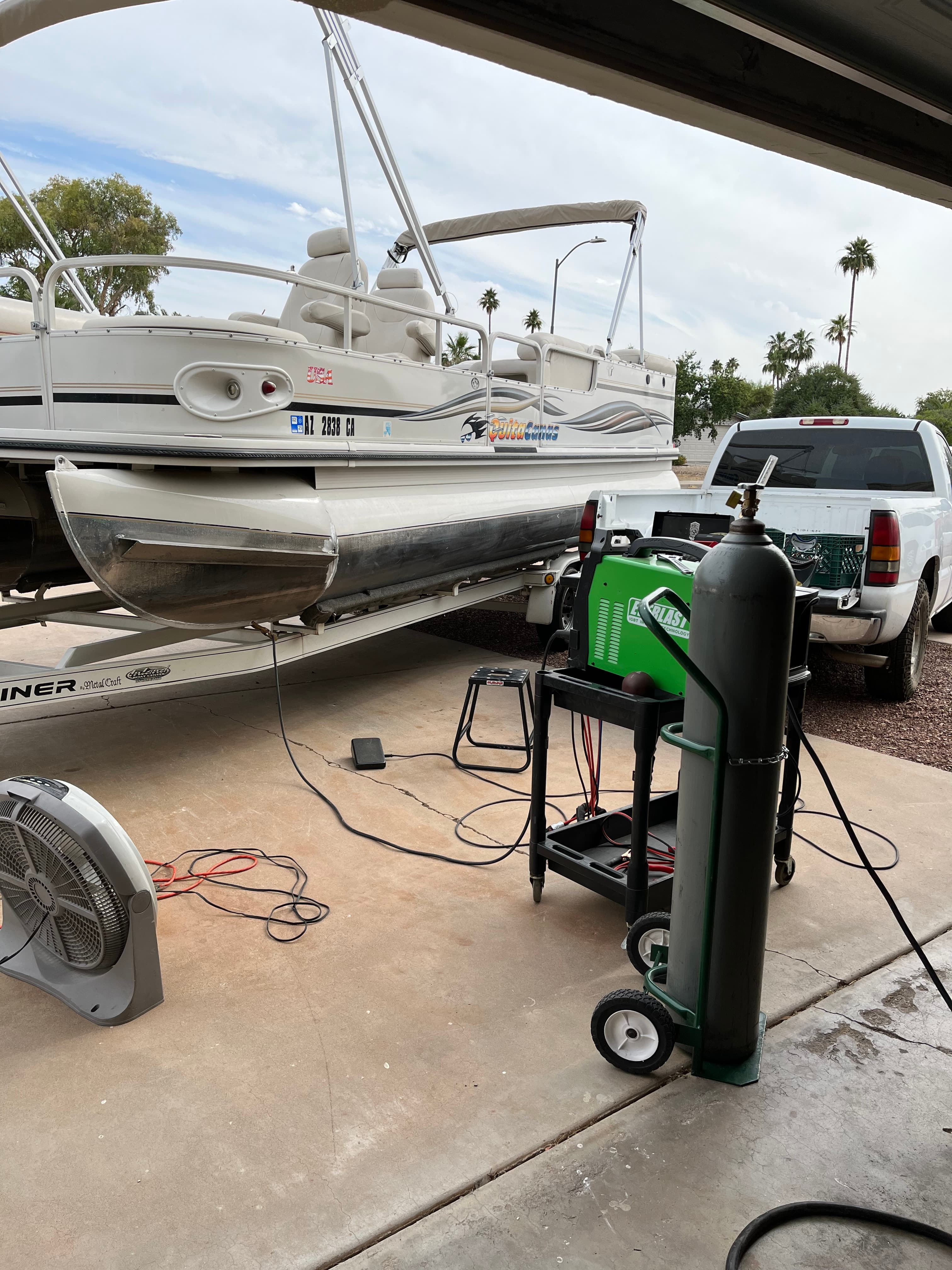 Pontoon boat on a trailer next to a welding machine and a white pickup truck.