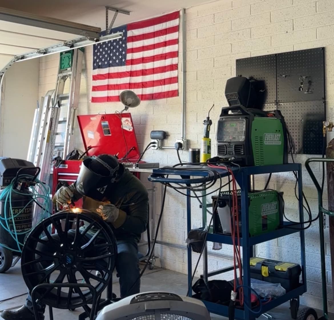 Welder in protective gear repairs a black car rim in a garage with American flag.