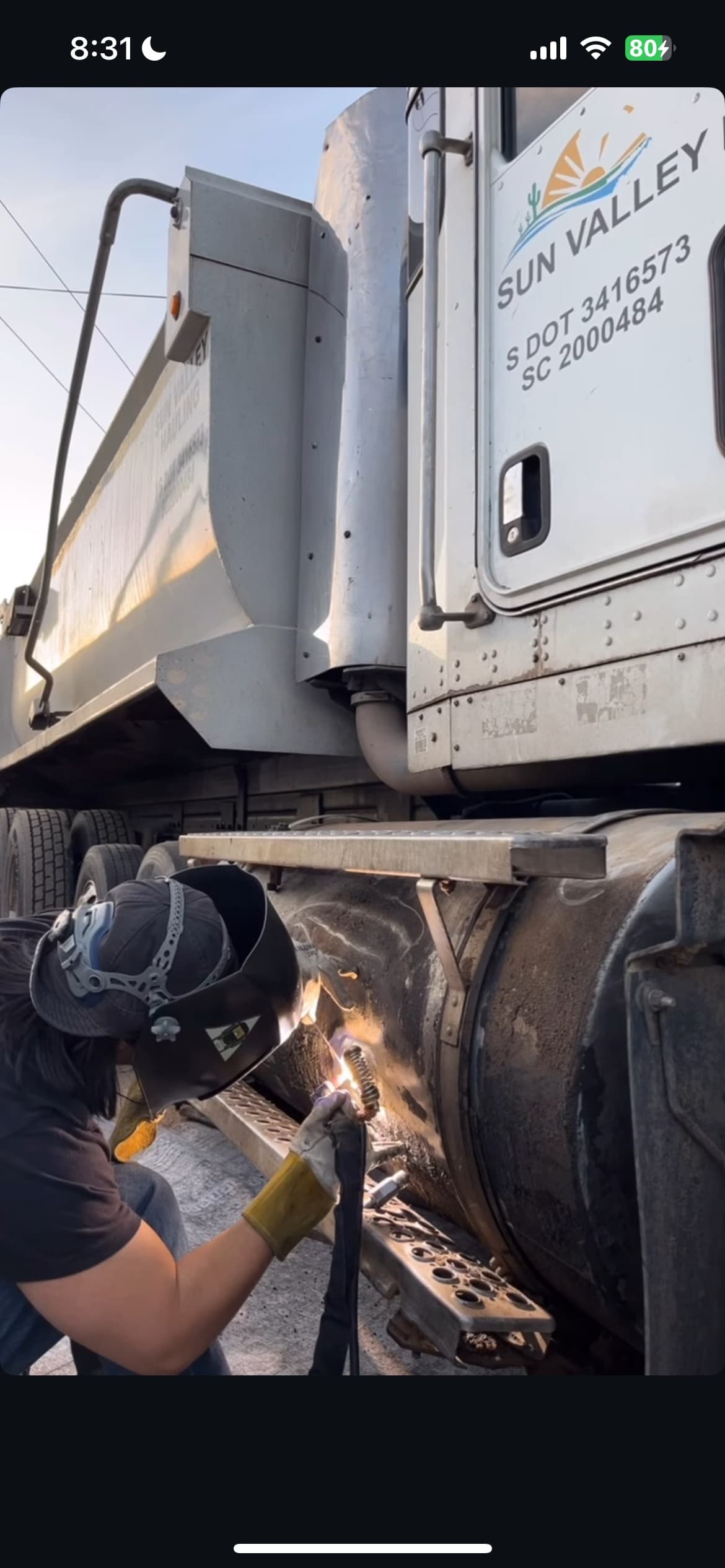 A welder in protective gear repairs a metal tank on a white commercial truck.